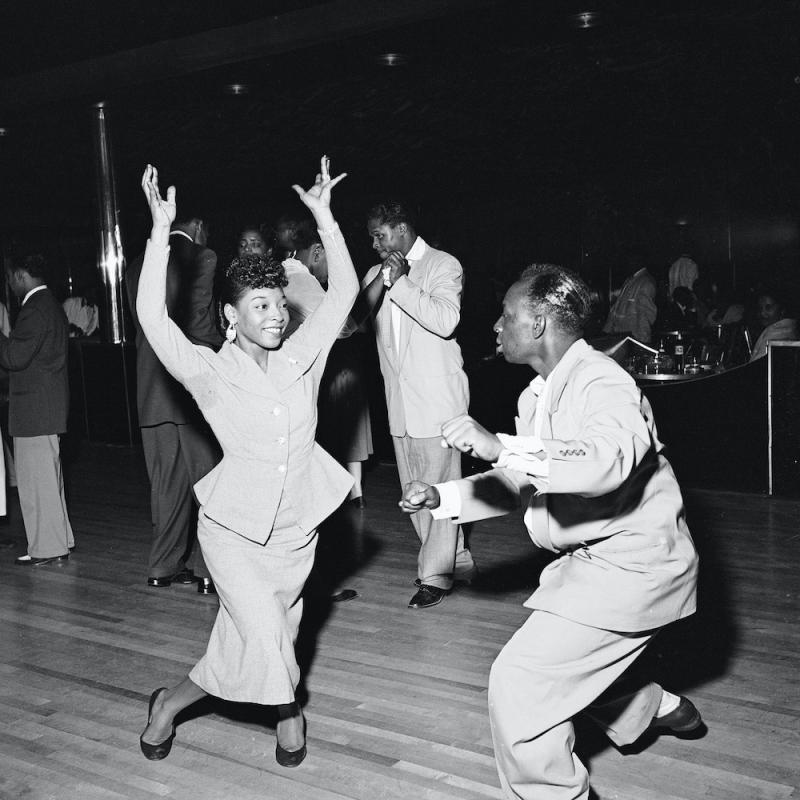 A Black woman smiles with her arms in the air as she dances with a Black man at the Savoy Ballroom in Harlem in New York City during the 1940s.
