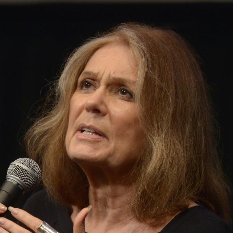 Author and feminist icon Gloria Steinem holds a microphone against a black backdrop